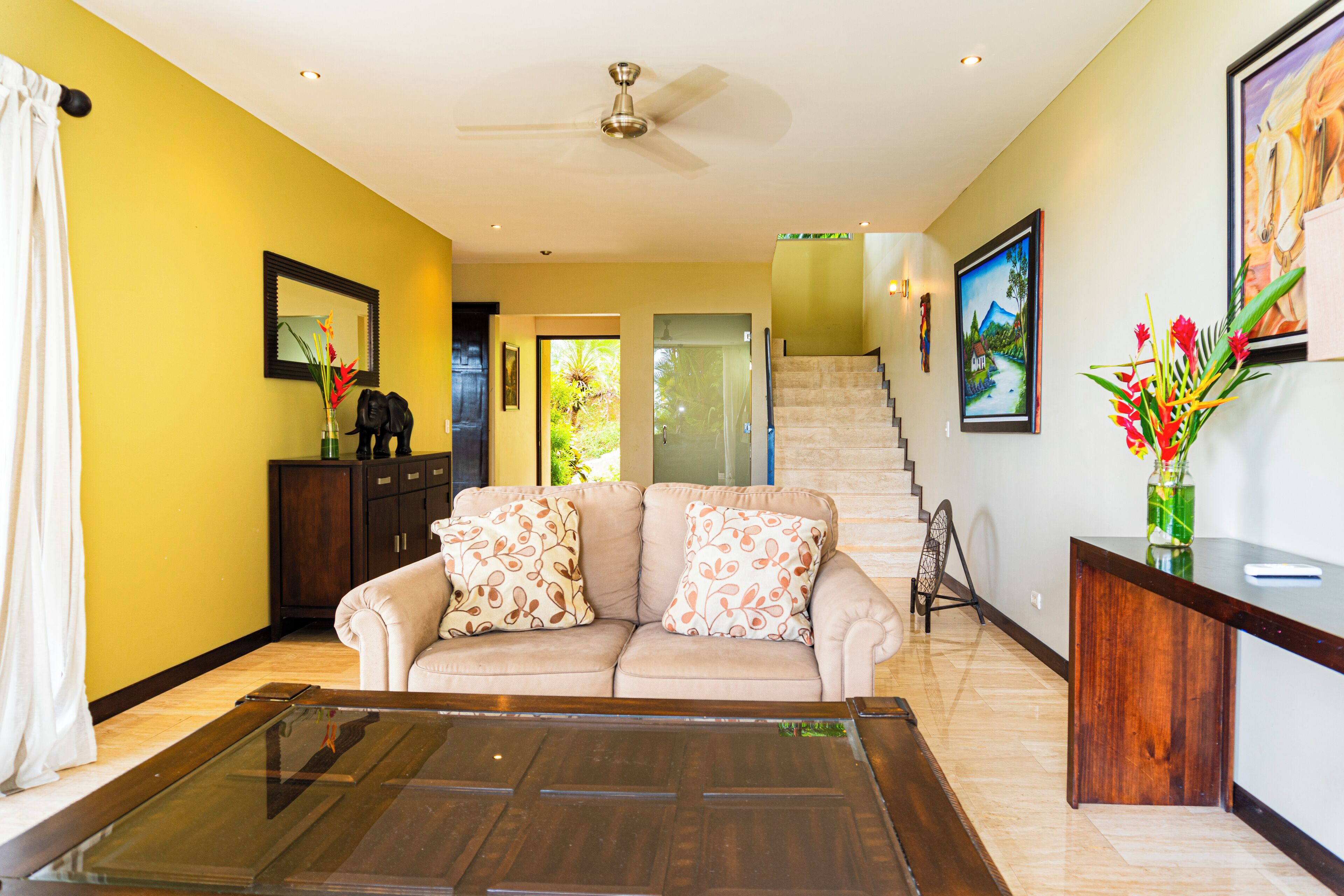 A living room with a beige sofa, a glass coffee table, and a wooden console table with a vase of flowers. A staircase with a glass railing leads to another room.