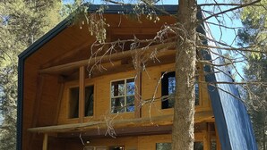 Una cabaña de troncos de dos pisos con un porche, escaleras de madera y un árbol grande al frente.