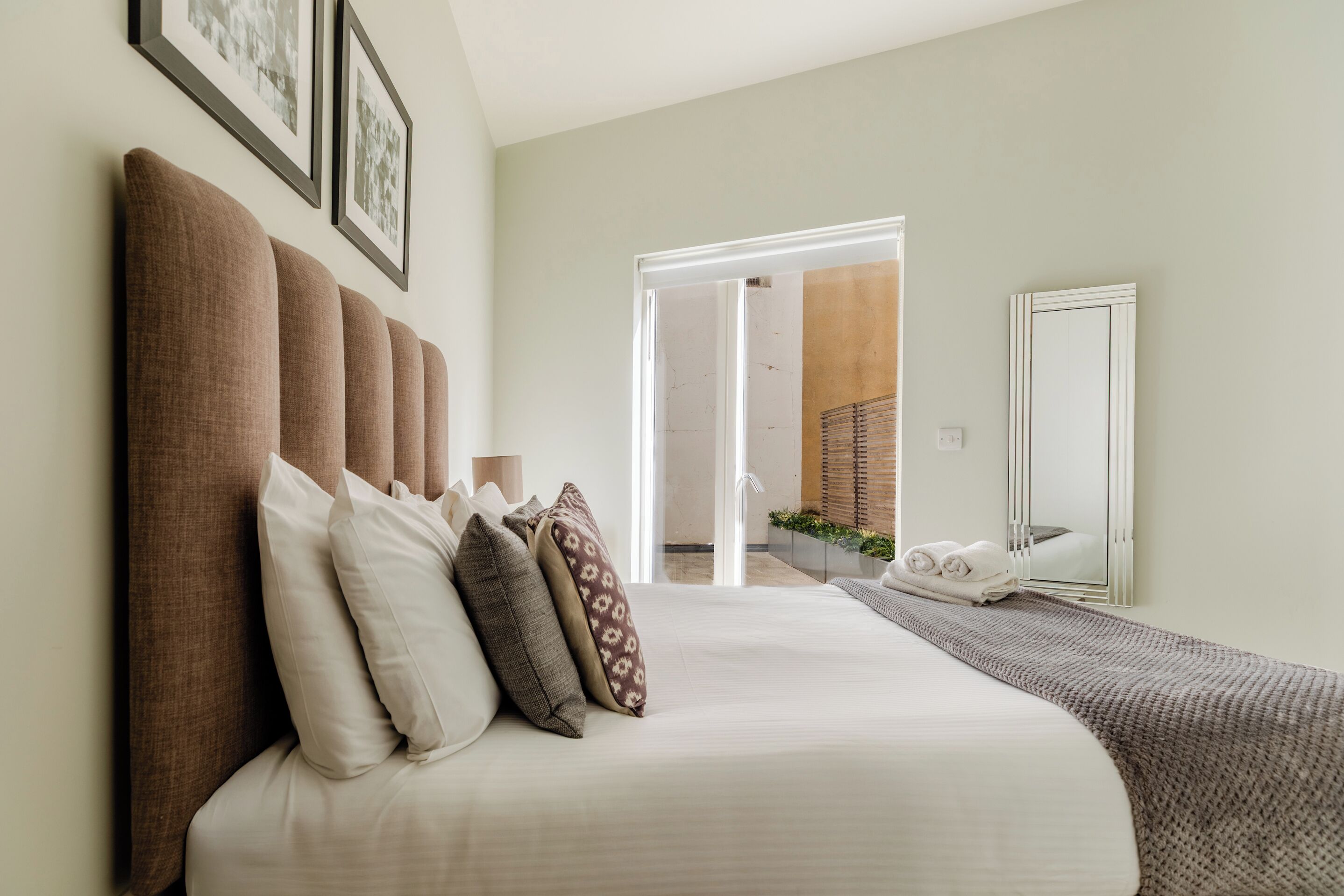 A neatly arranged bedroom with a large bed, multiple pillows, a bedside table, and a door leading to an outdoor area.