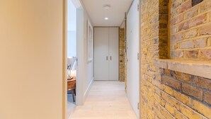A narrow hallway with a wooden floor, a brick wall on one side, and a white door at the end. - Excel Apartments Ltd (Bromley)