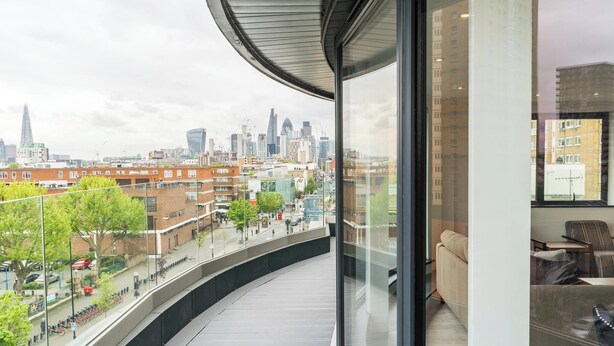 A modern apartment with a curved balcony overlooking a cityscape, featuring a view of the skyline and a street with bicycles. - Excel Apartments Ltd (Bromley)