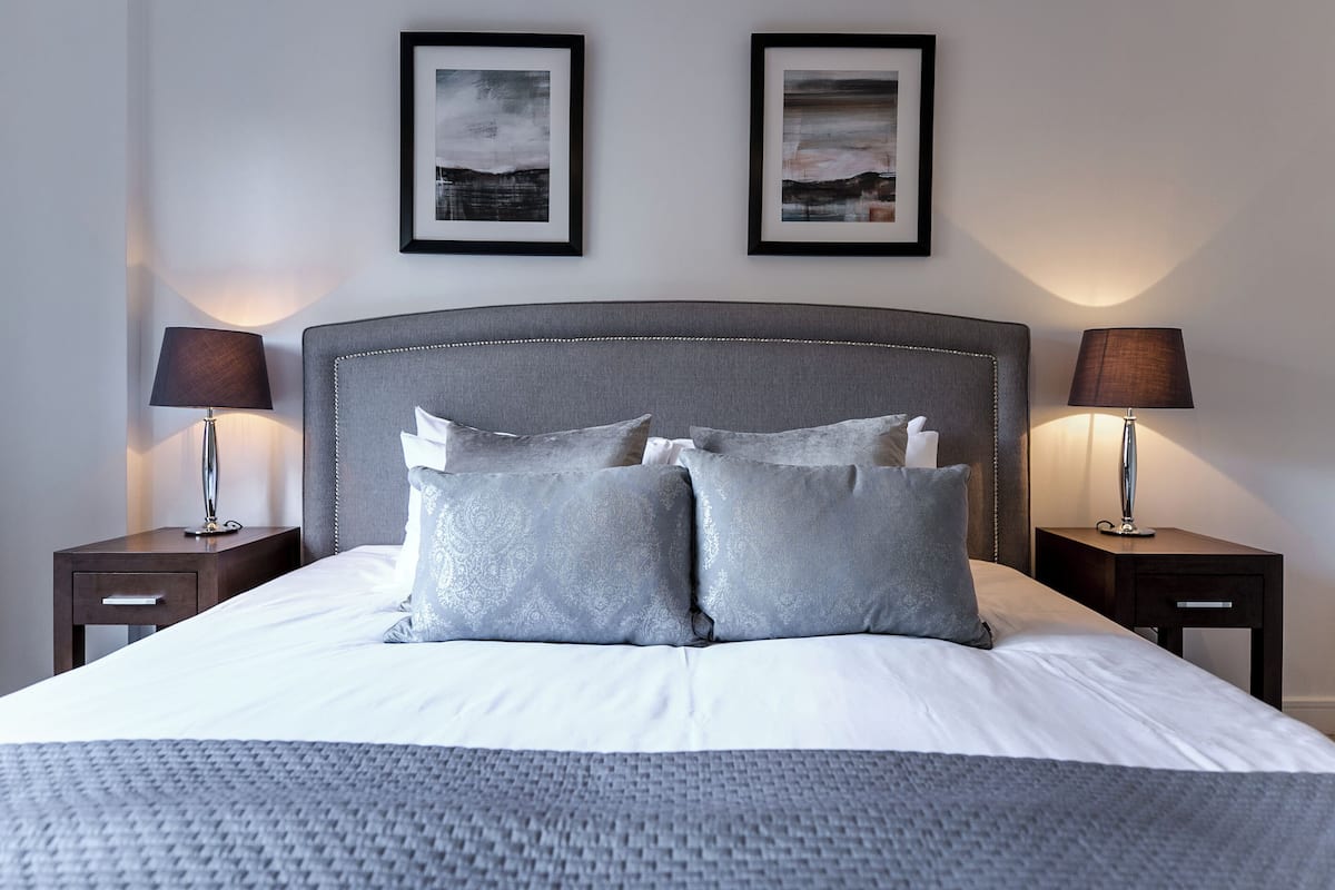 A neatly made bed with a grey headboard, two bedside tables with lamps, and two framed pictures on the wall.