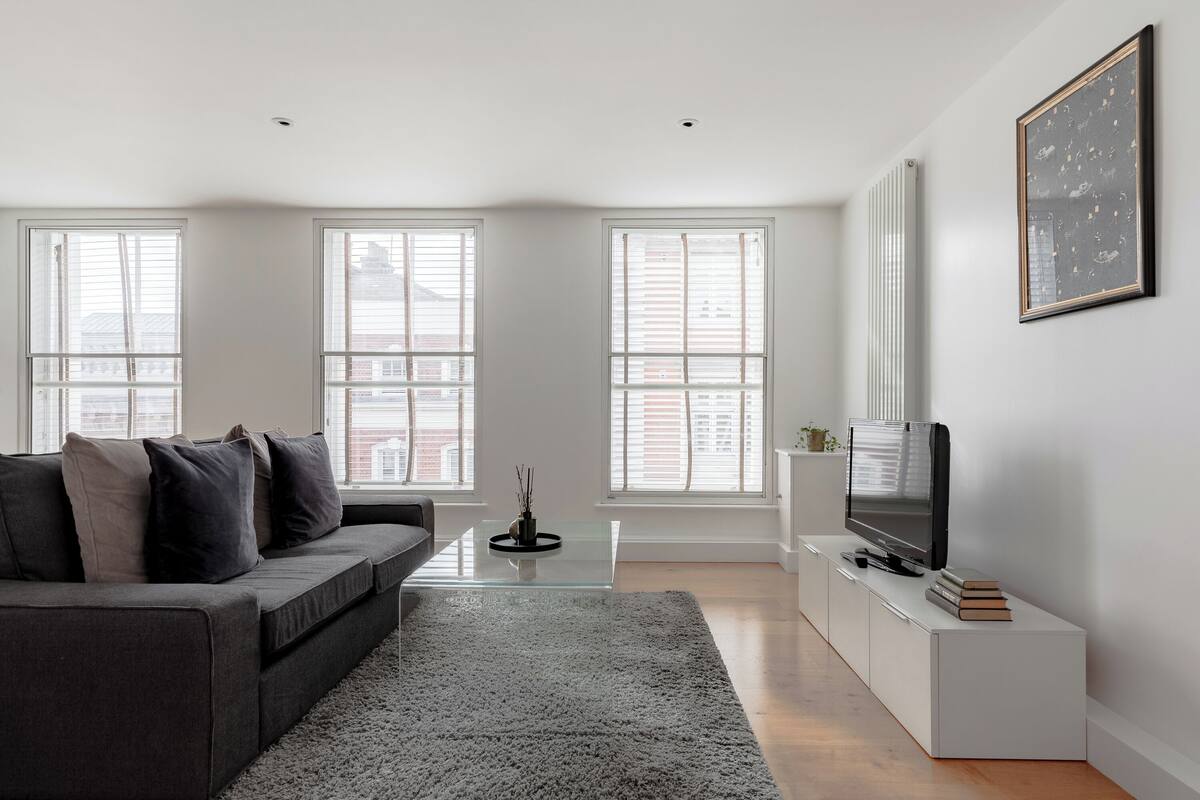 A modern living room with a grey sofa, a glass coffee table, a flat-screen TV on a white stand, and large windows with blinds.