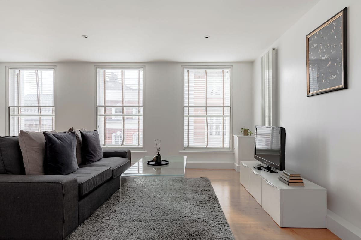 A modern living room with a grey sofa, a glass coffee table, a flat-screen TV on a white stand, and large windows with blinds.