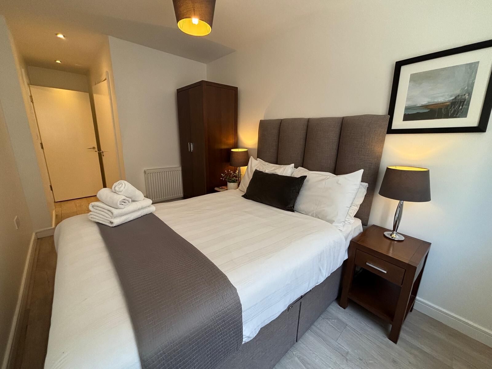 A neatly made bed with a grey headboard, a bedside table with a lamp, and a framed picture on the wall.