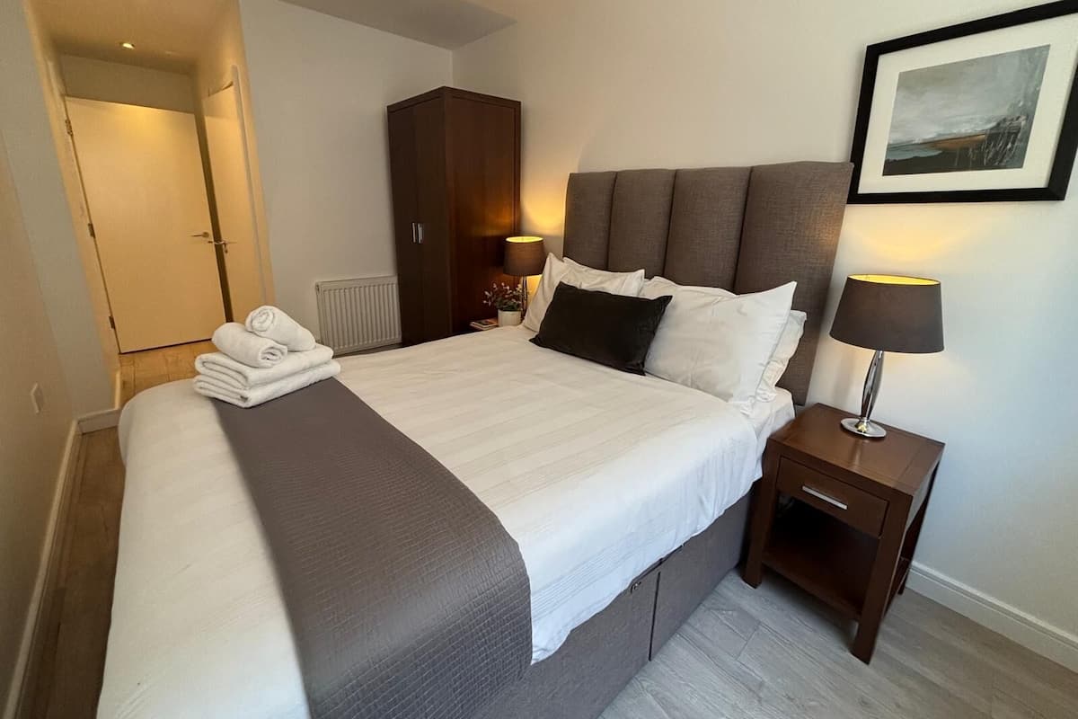 A neatly made bed with a grey headboard, two bedside lamps, a framed picture on the wall, and a door leading to another room.
