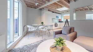 A modern living room with a glass dining table, white chairs, a grey sofa, and a kitchen in the background. - Excel Apartments Ltd (Bromley)