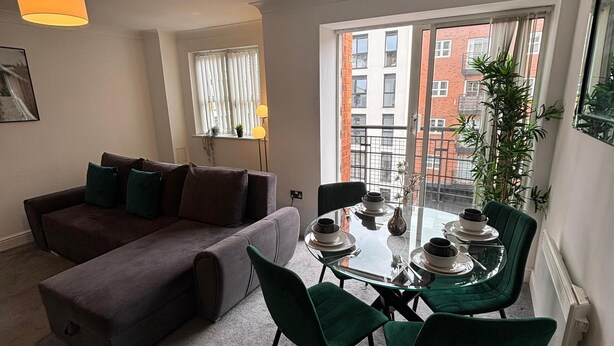 Una sala de estar moderna con una mesa de comedor de vidrio, sillas verdes, un sofá gris y un ventanal con vistas a edificios.