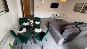 Una sala moderna con una mesa de comedor de vidrio preparada para cuatro personas, rodeada de sillas de terciopelo verde. Se ve un sofá gris y una pequeña mesita de noche con una lámpara.