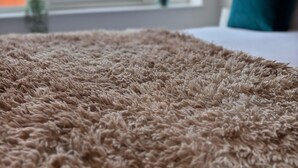 Primer plano de una alfombra beige con textura en una habitación con un gran ventanal, una cama con ropa de cama blanca y una almohada color turquesa.