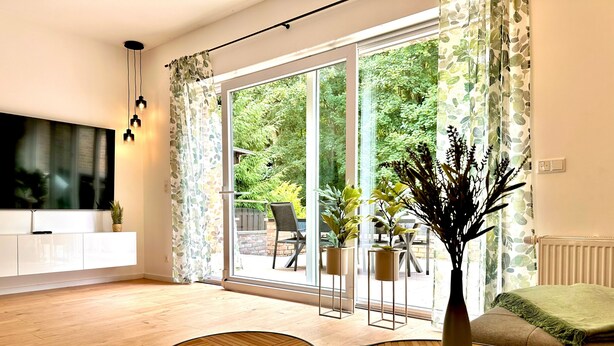Una sala de estar moderna con un gran ventanal corredero que da a un patio exterior, un televisor de pantalla plana montado en un mueble blanco, dos mesas redondas de madera y una planta en maceta sobre una mesita auxiliar.