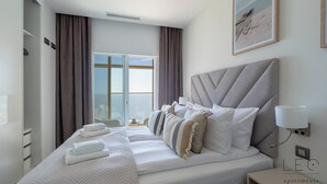 Un dormitorio moderno con una cama grande, una mesita de noche, una lámpara de pared y vistas al exterior a través de una ventana.
