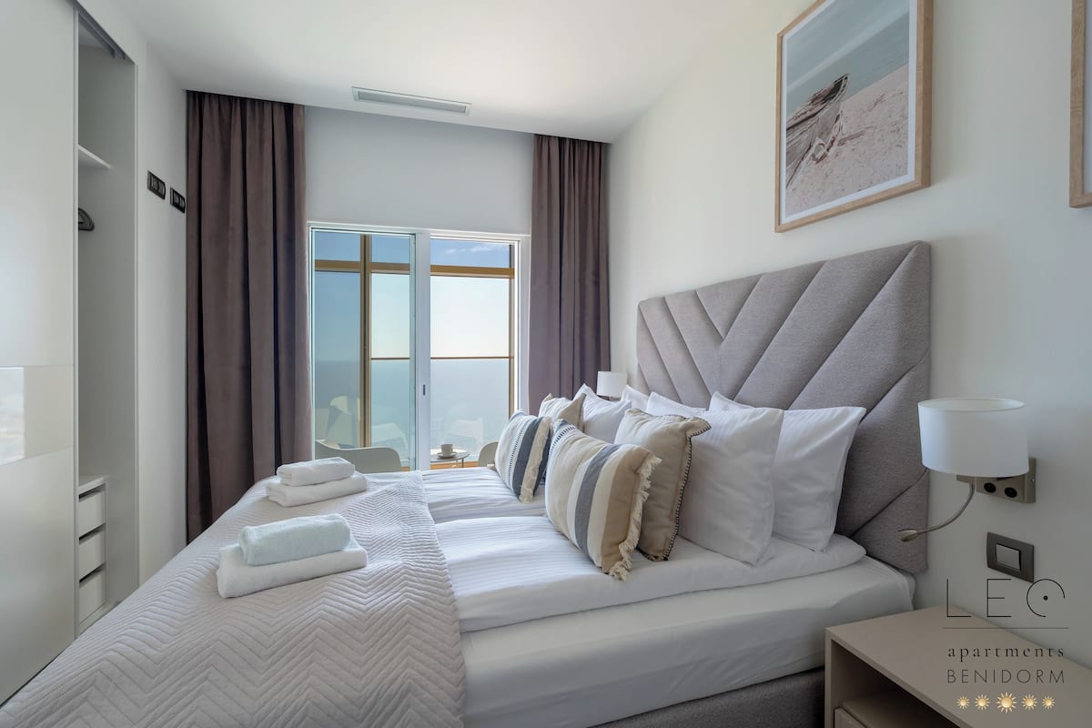 Ein modernes Schlafzimmer mit einem großen Bett, einem Nachttisch, einer Wandleuchte und Blick ins Freie durch ein Fenster.