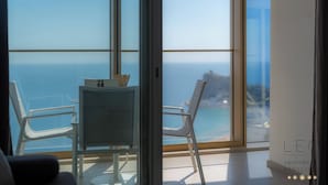 Una sala moderna con un gran ventanal que da a un balcón con vistas al mar. Hay dos sillones blancos y una mesita con un jarrón.