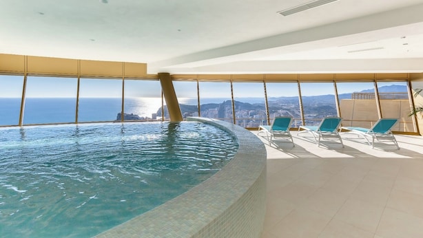 Una piscina infinita en el interior con vista a la ciudad, sillas de descanso y suelo de baldosas.