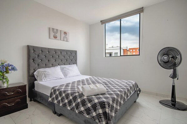 A bedroom with a grey upholstered bed, white pillows, a checkered blanket, a bedside table with a vase of flowers, and a window with a city view.