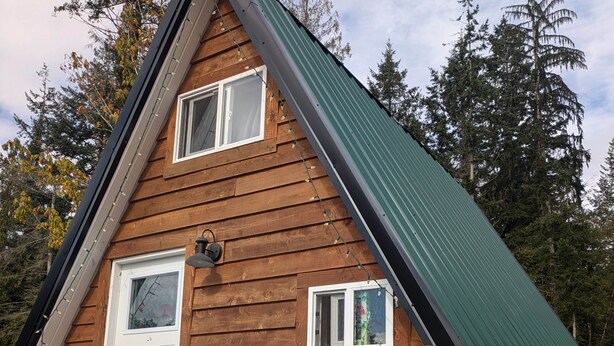 Une cabane en bois avec un toit vert, deux fenêtres, une porte et un petit porche avec des chaises bleues.
