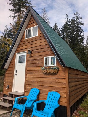 Eine Holzhütte mit grünem Dach, zwei Fenstern, einer Tür und einer kleinen Veranda mit blauen Stühlen.