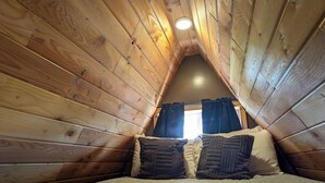 Une chambre confortable située dans les combles, avec des murs en bois, un plafond en pente, une fenêtre unique agrémentée de rideaux et un lit avec deux oreillers.
