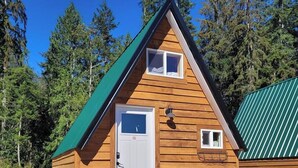 Une petite cabane en bois avec un toit vert, une porte blanche et deux chaises Adirondack rouges à l’extérieur.