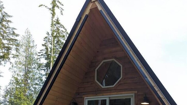 A wooden cabin with a pitched roof, a large sliding glass door, and two blue Adirondack chairs on the porch. - Golden rings cabin on Homestead W/Hot Tub (qathet Regional District)