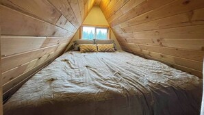 A small, wooden-paneled attic bedroom with a sloped ceiling, a single bed, and a window providing a view of trees. - Golden rings cabin on Homestead W/Hot Tub (qathet Regional District)