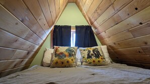 Pequeño dormitorio en ático, de forma triangular, con paredes de madera, una cama con dos almohadas estampadas y un ventanal con cortinas negras.