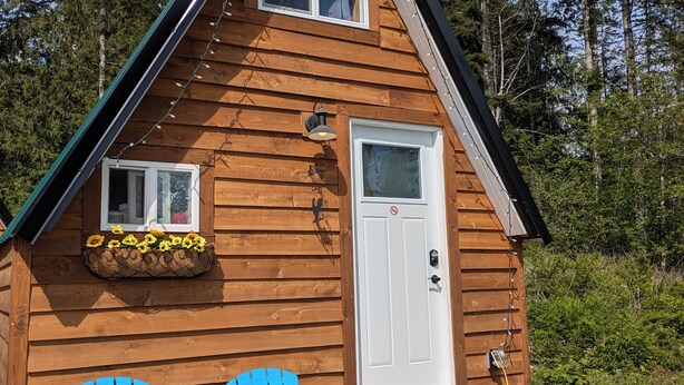 Una cabaña de madera con dos sillas Adirondack azules en la entrada, una puerta blanca y un ventanal pequeño.