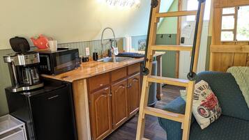 A compact kitchen with a coffee maker, microwave, and wooden cabinets. A ladder is leaning against the wall, and a couch with a pillow is visible in the background.