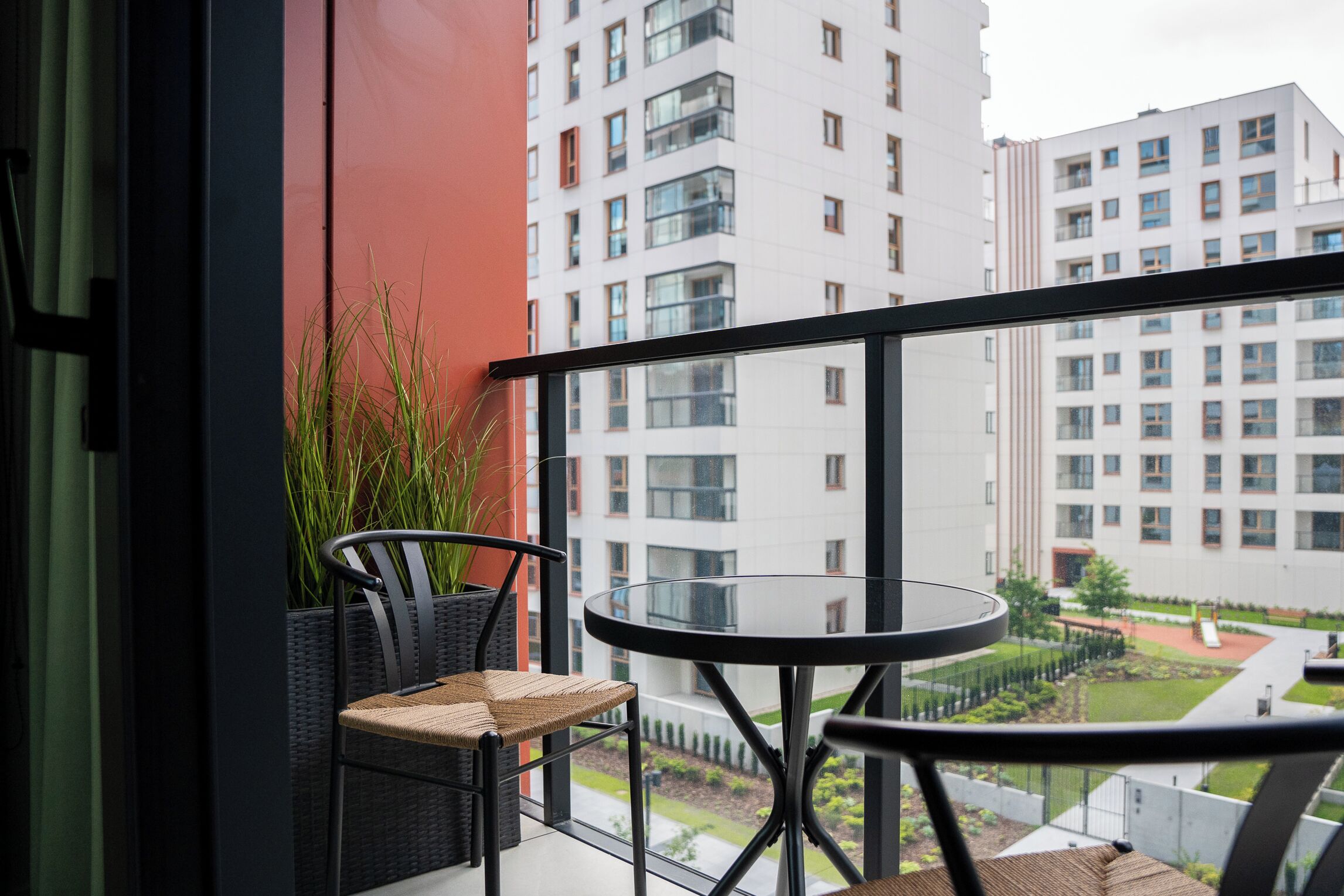 A balcony with a round table and two wicker chairs, overlooking a residential area with modern buildings.