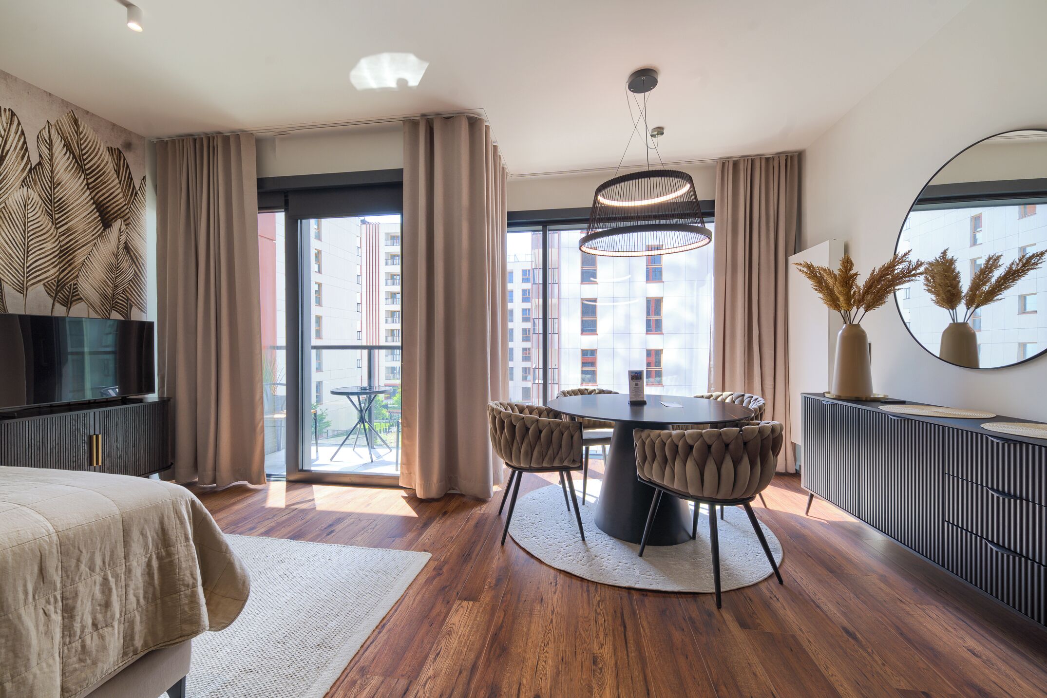 A modern hotel room with a large bed, a dining area with a round table and chairs, a TV, and a view of the city through large windows.