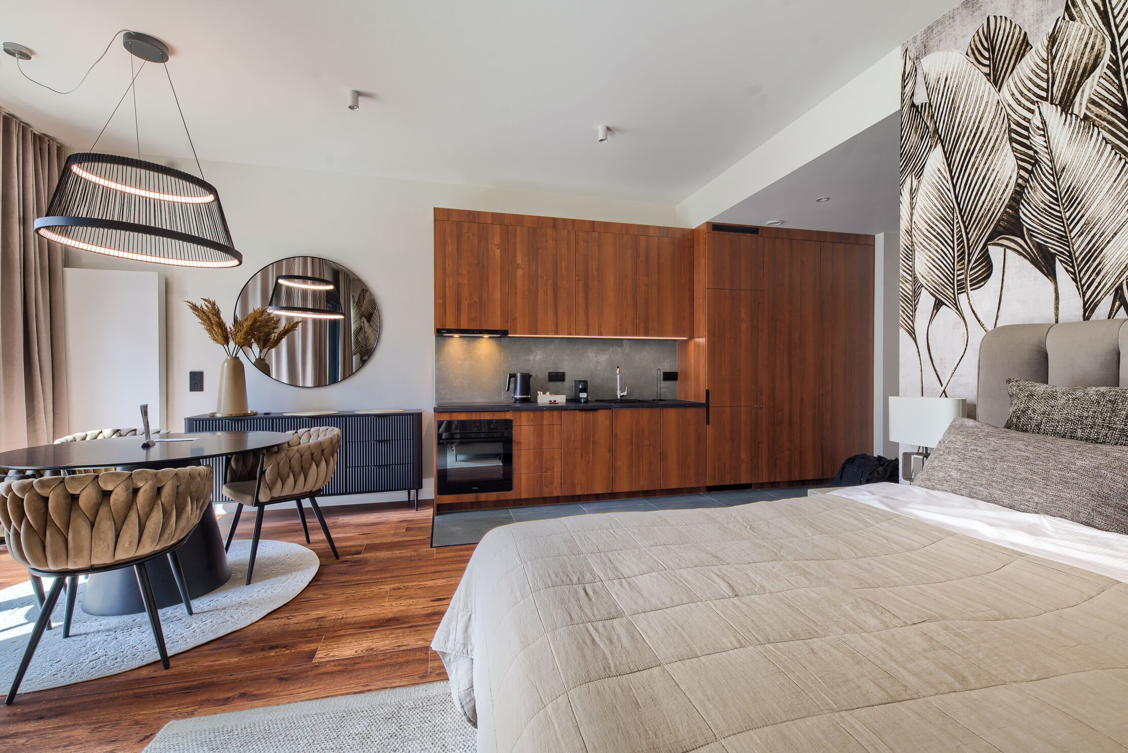 A modern bedroom with a large bed, a dining area with a round table and chairs, and a kitchenette with wooden cabinets.