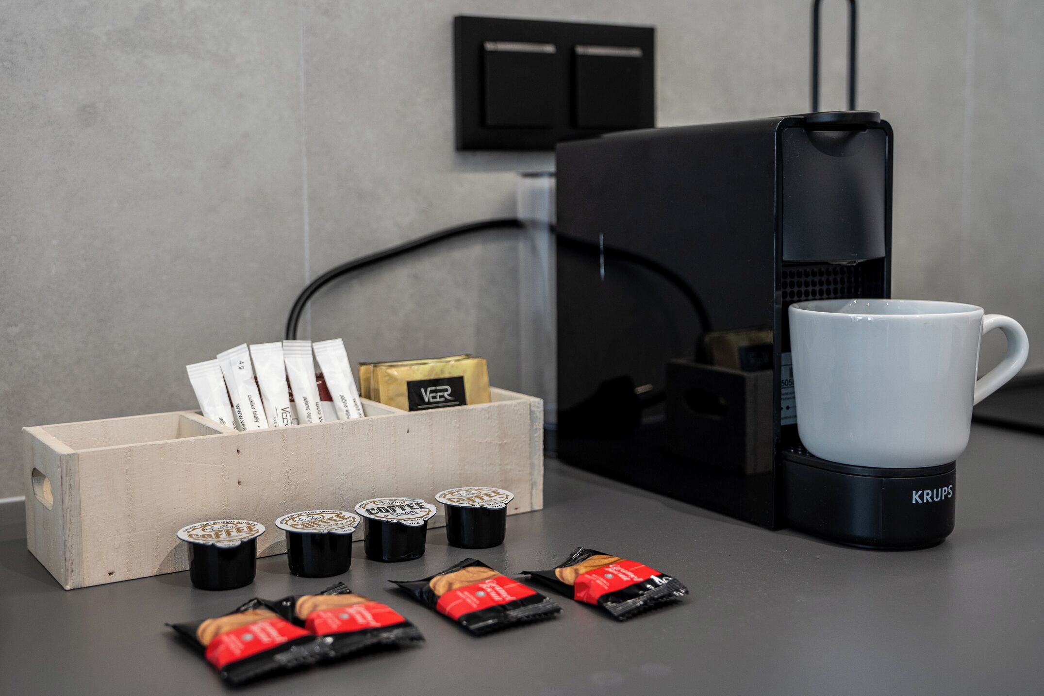 A coffee maker, a white mug, and a wooden box with coffee pods and packets on a countertop.