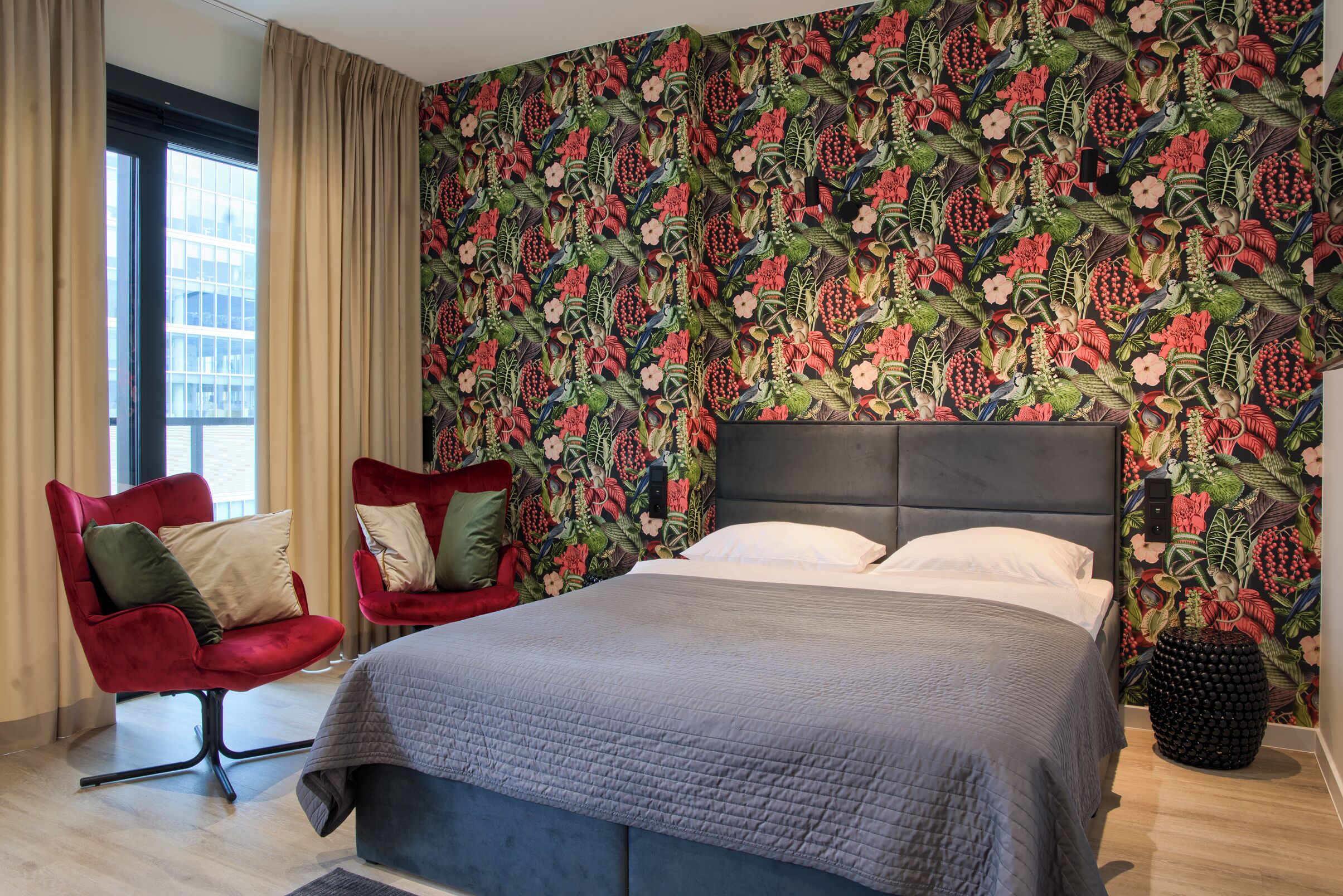 Un dormitorio con papel tapiz floral, una cama con colcha gris, dos sillas rojas y una mesita de noche negra.