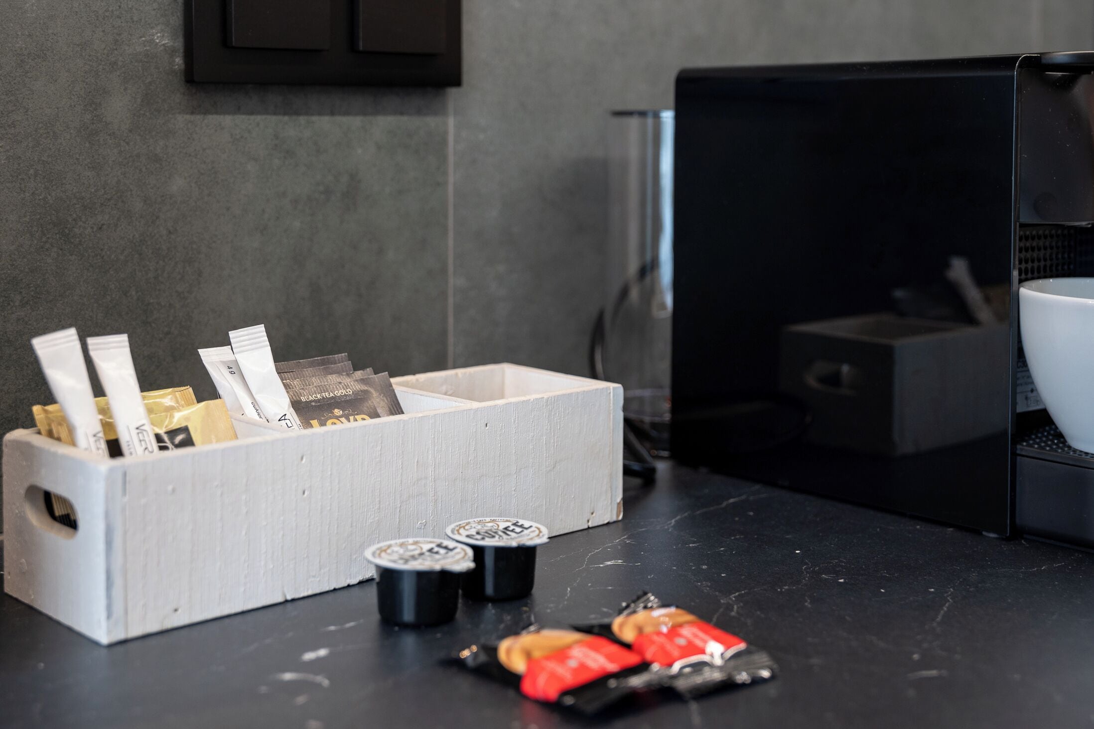 Un comptoir de cuisine avec une boîte de rangement pour le café contenant des dosettes de café et quelques sachets.