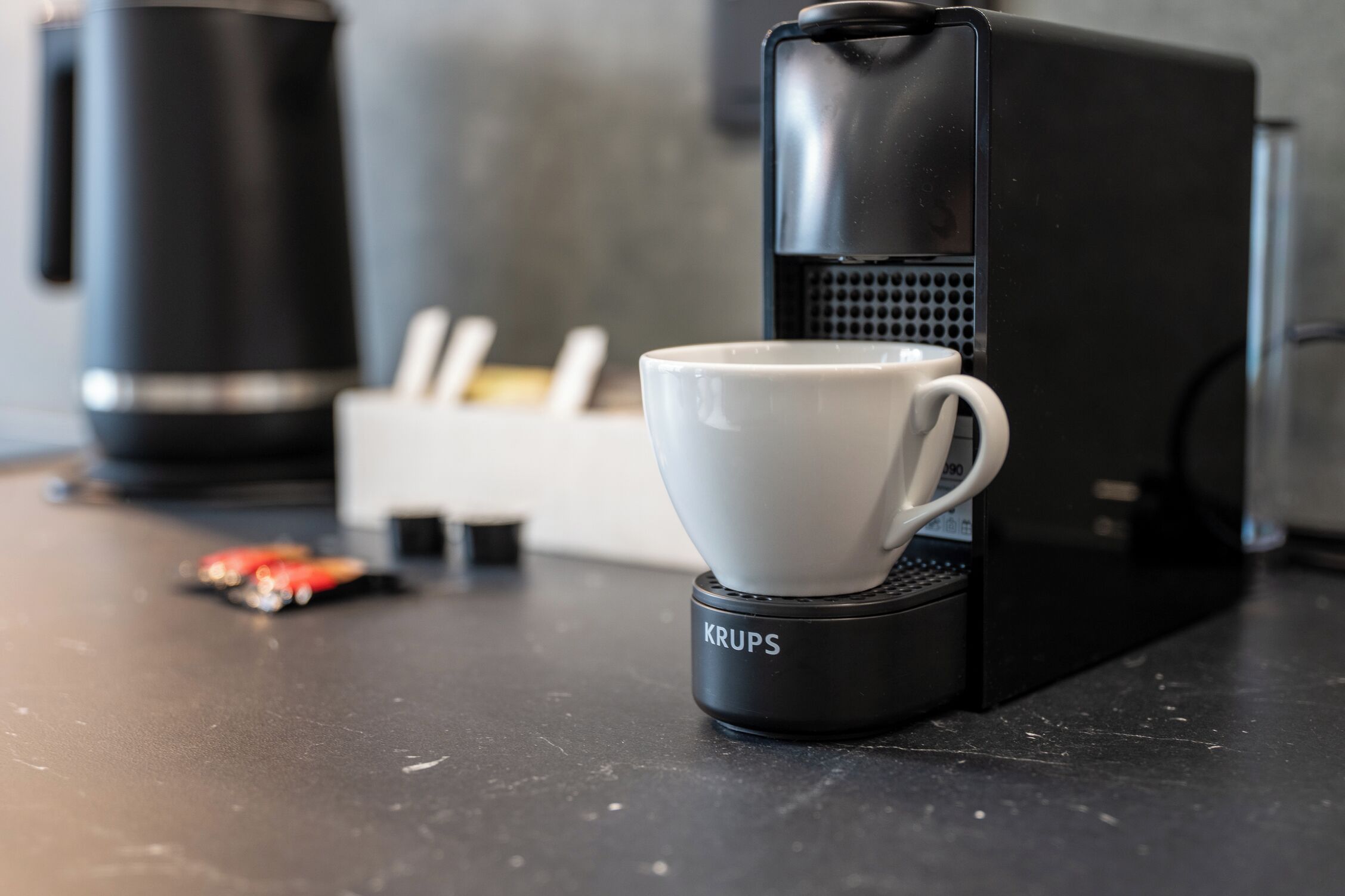 A coffee maker with a cup on its base, a coffee pot, and a container with utensils on a countertop.