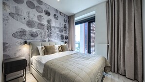 A modern bedroom with a large bed, a nightstand, a wall mural, and a view through a window with curtains. - Towarowa Towers (Warsaw)