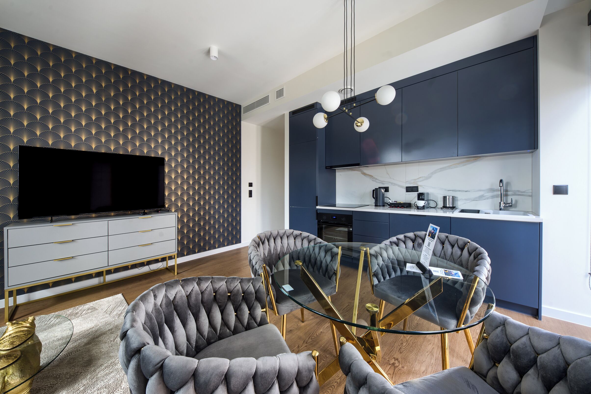 Un salon moderne avec une table basse en verre, des fauteuils à capitons, un téléviseur à écran plat et une cuisine bleu foncé aux meubles blancs.
