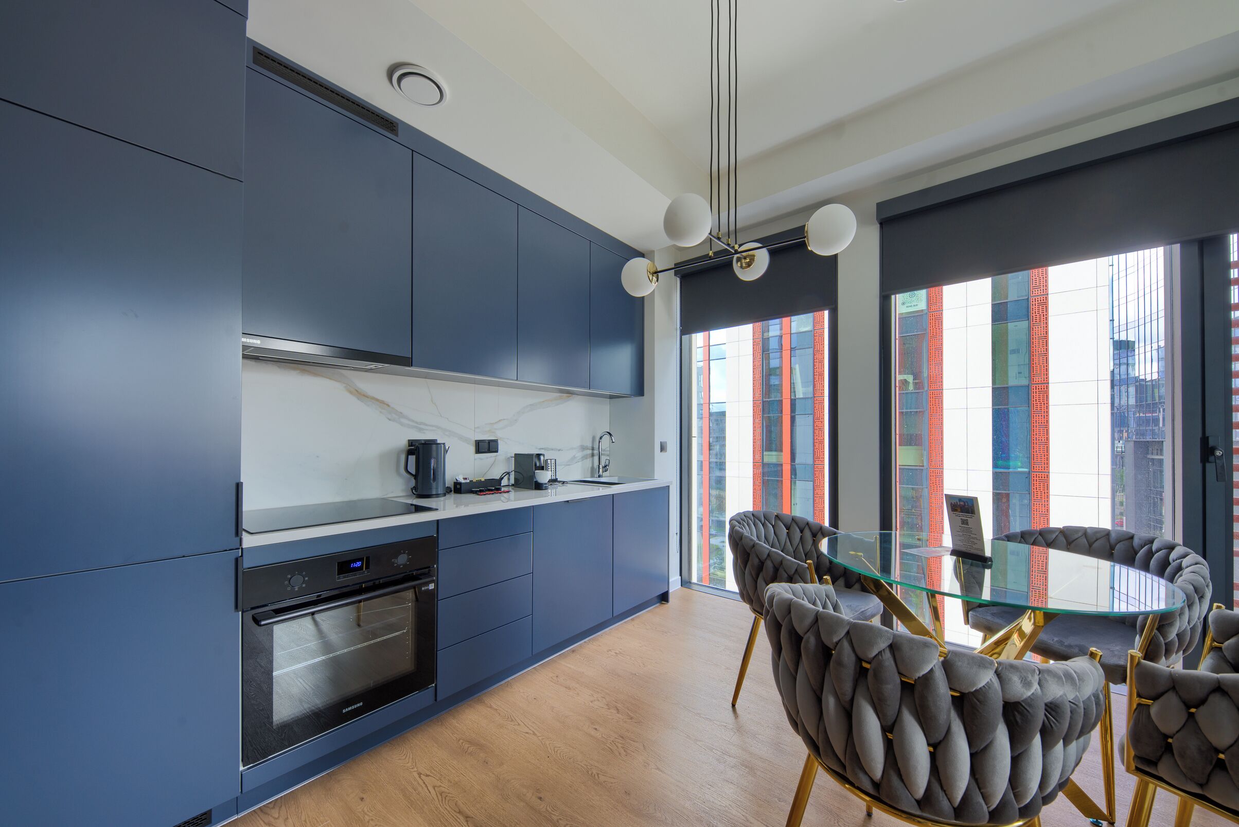 A modern kitchen with built-in appliances, a dining area with a glass table and cushioned chairs, and a city view through large windows.