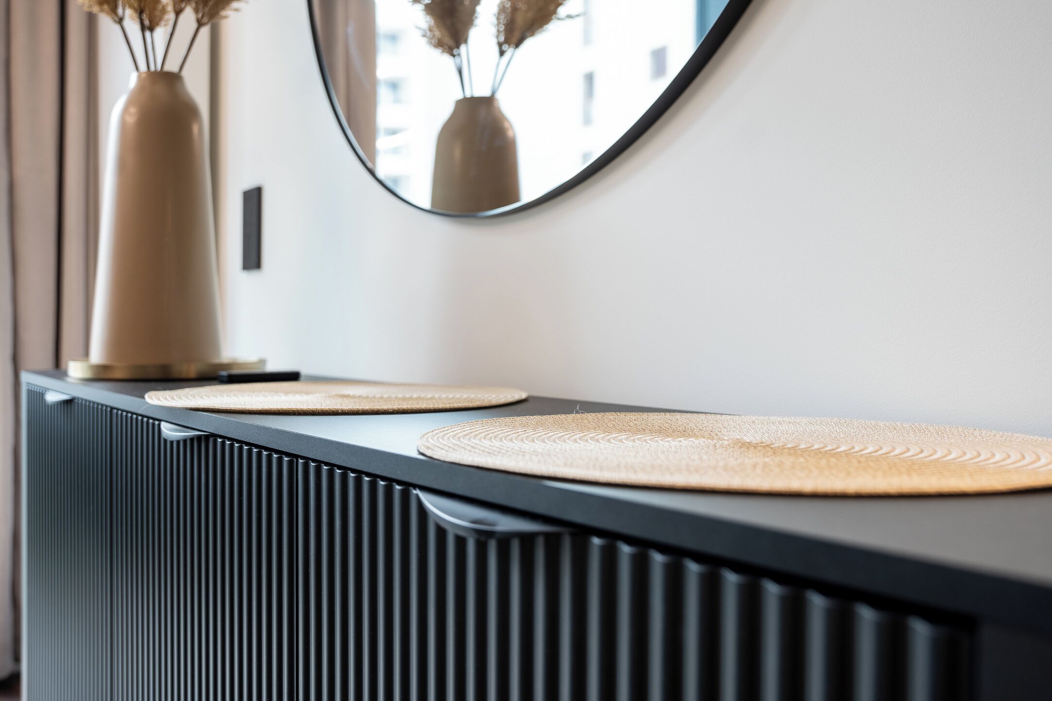 A modern bathroom vanity with a black countertop, two woven placemats, a tall vase with dried flowers, and a mirror reflecting a building.