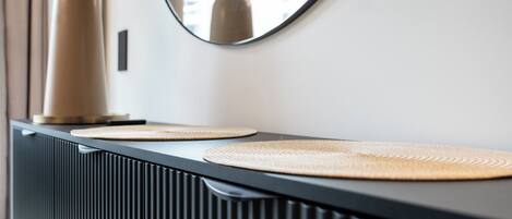 A modern bathroom vanity with a black countertop, two woven placemats, a tall vase with dried flowers, and a mirror reflecting a building.