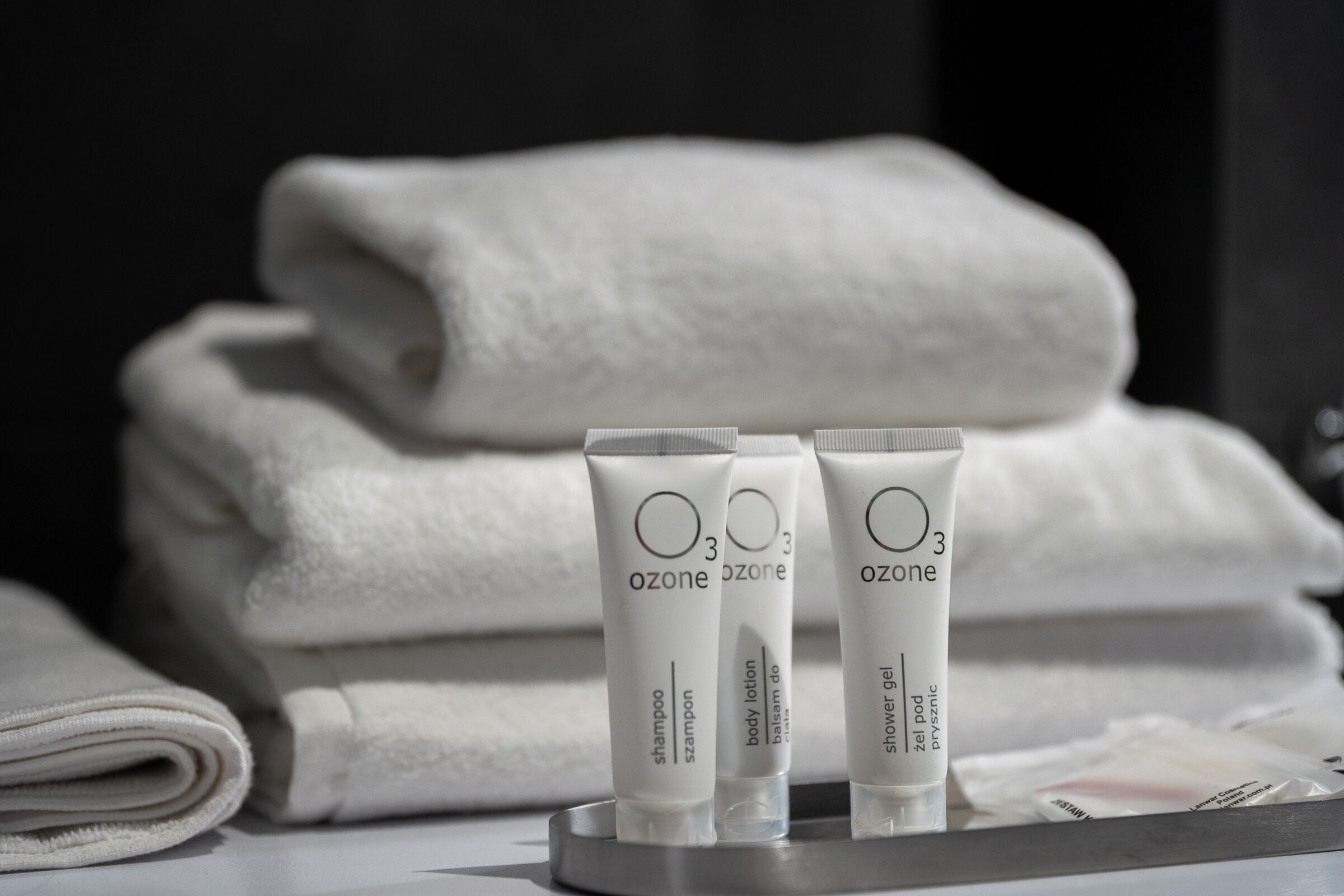 A bathroom with neatly folded white towels stacked in the background and three tubes of Ozone body lotion on a tray in the foreground.