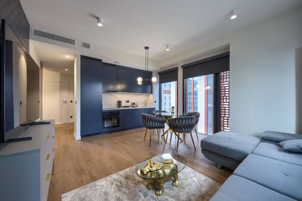 Un salon moderne avec un canapé gris, une table basse en verre agrémentée d’un objet décoratif, un coin repas avec une table et des chaises, et une cuisine avec des meubles bleu foncé et un plan de travail blanc.