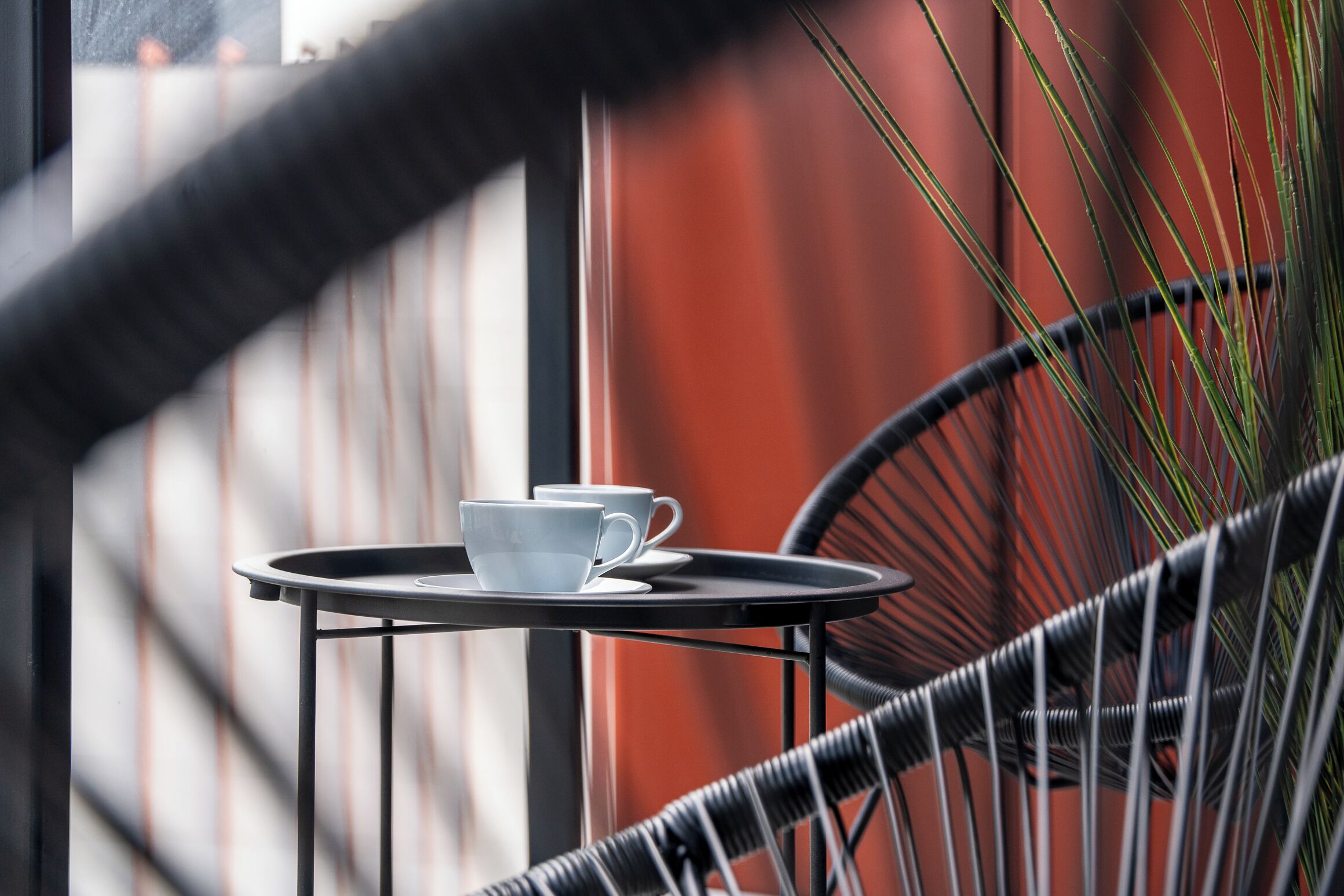 A small round table with a white cup and saucer on it, positioned on a balcony with a wicker chair and a potted plant in the background.