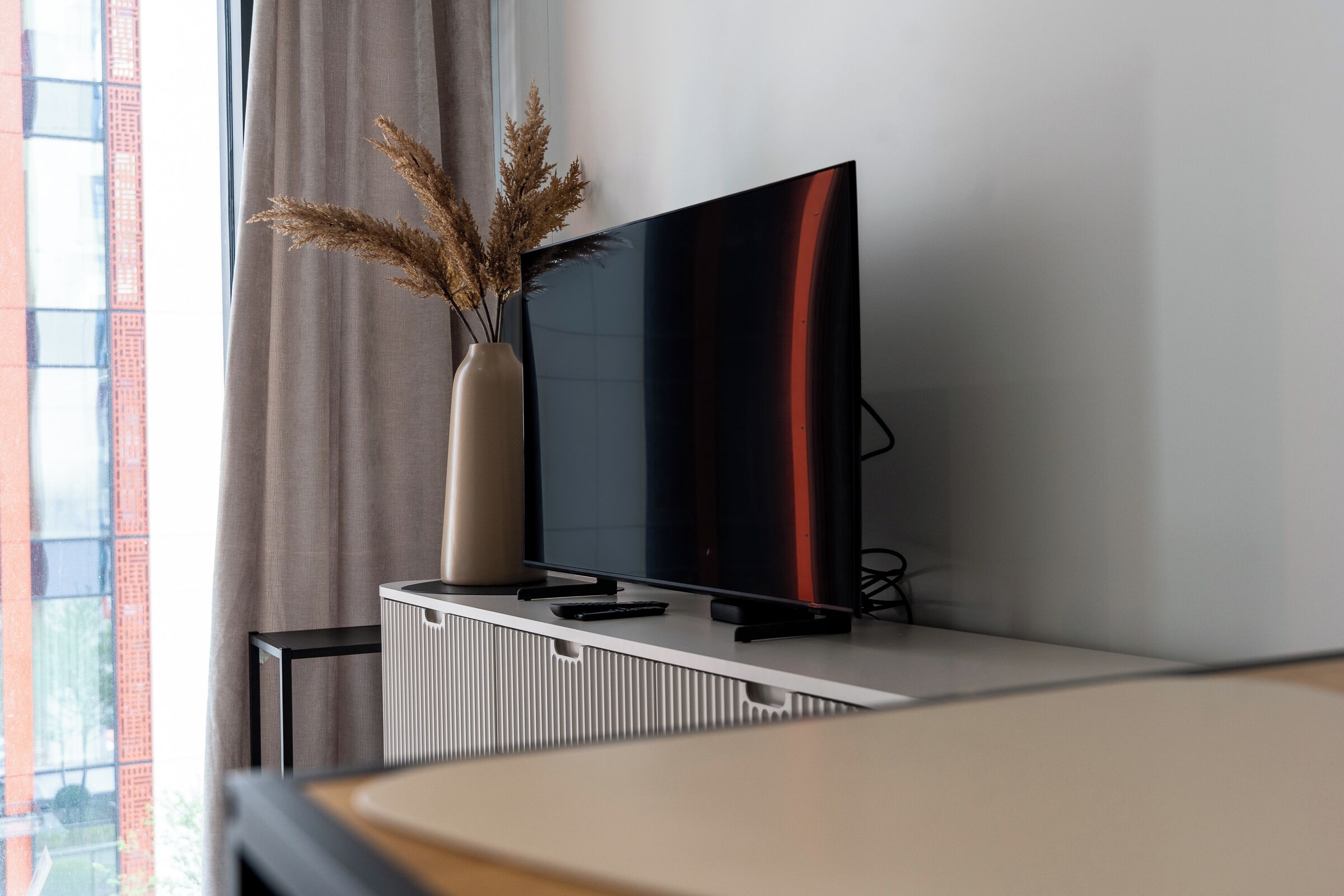 A modern living room with a flat-screen TV on a white console, a vase with dried plants, and a window with a view of a red building.
