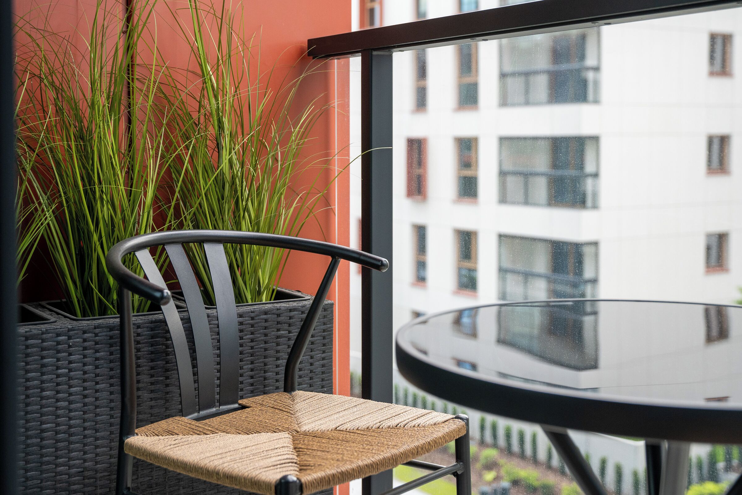 A wicker chair with a cushioned seat and backrest, positioned next to a glass table. A tall potted plant with long green leaves is visible in the background.