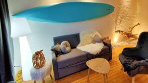 A modern living room with a grey sofa, a round wooden coffee table, and a black armchair. A large blue surfboard decorates the wall, and a rustic wooden sculpture is on the floor. - Balneo 100m beach (Berck)