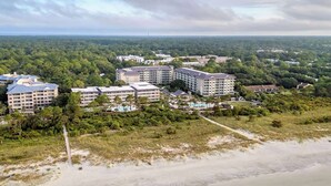 Ein Hotelkomplex an der Küste mit mehreren Gebäuden, einem Strand im Vordergrund und bewaldetem Gelände im Hintergrund.