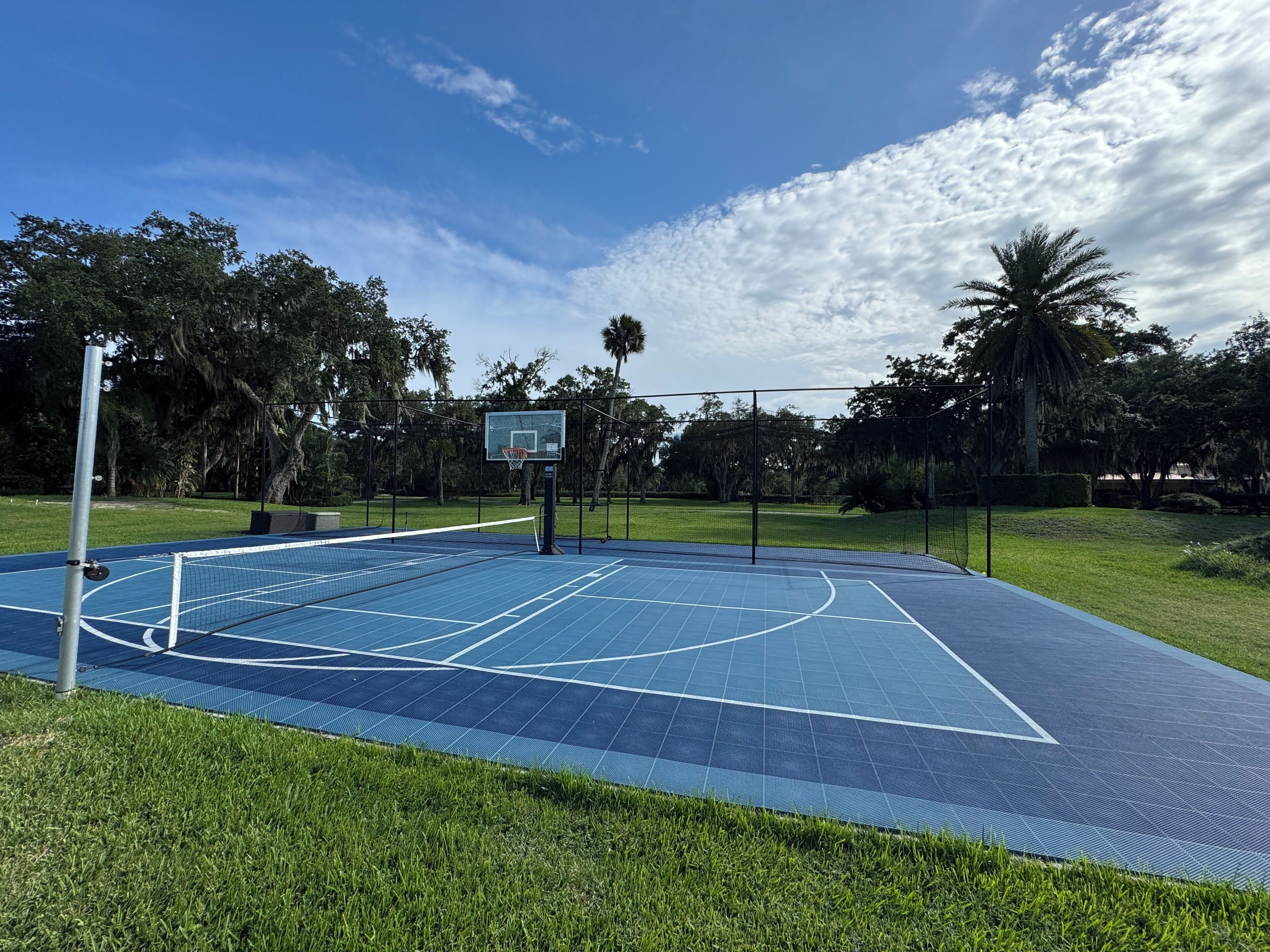 An outdoor basketball court with a blue surface, surrounded by grass and trees.