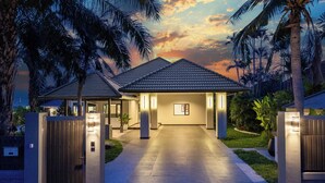 A modern house with a paved driveway leading to a covered entrance, surrounded by palm trees and a sunset sky.